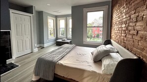 A bedroom with a large bed, a desk, and a view of a red building through the windows.