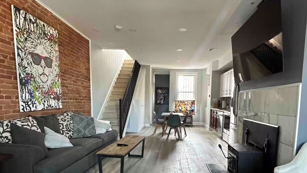 A modern living room with a grey sofa, a coffee table, and a fireplace. A staircase leads to a dining area with a table and chairs.