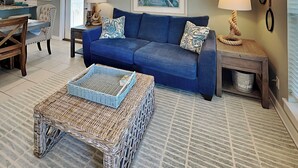 Un salon avec un canapé bleu, une table basse en osier, un tableau accroché au mur et, au fond, un coin cuisine.