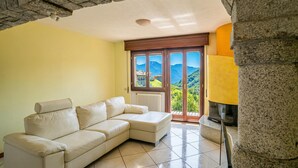 A living room with a beige sectional sofa, a stone column, a fireplace, and a view of mountains through a large window. - Balcone Di Alex & Thomas, Val Rezzo, Italy (Val Rezzo)