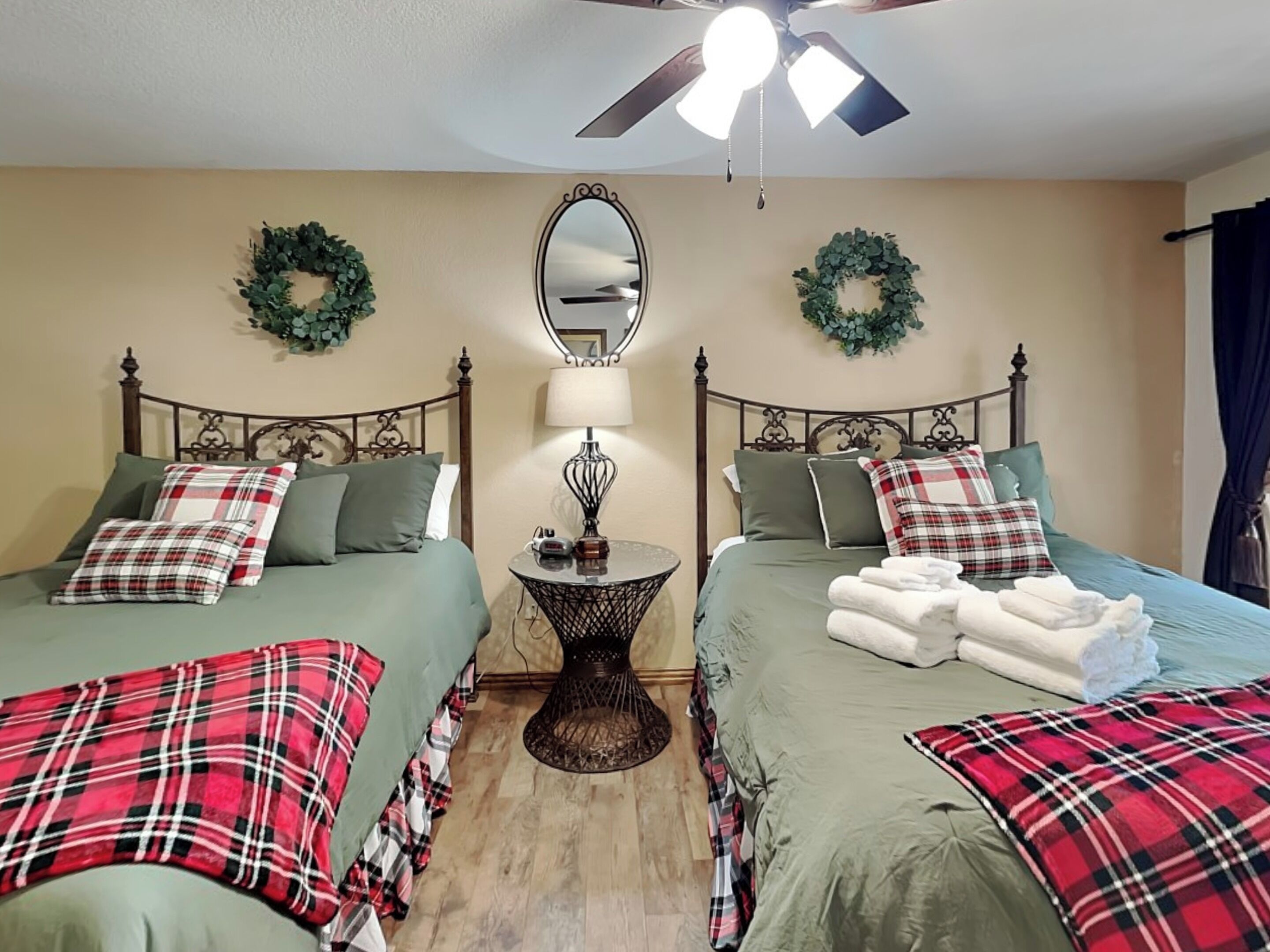 A bedroom with two beds, each with green bedding and plaid pillows, a round table with a lamp, and two wall-mounted wreaths.