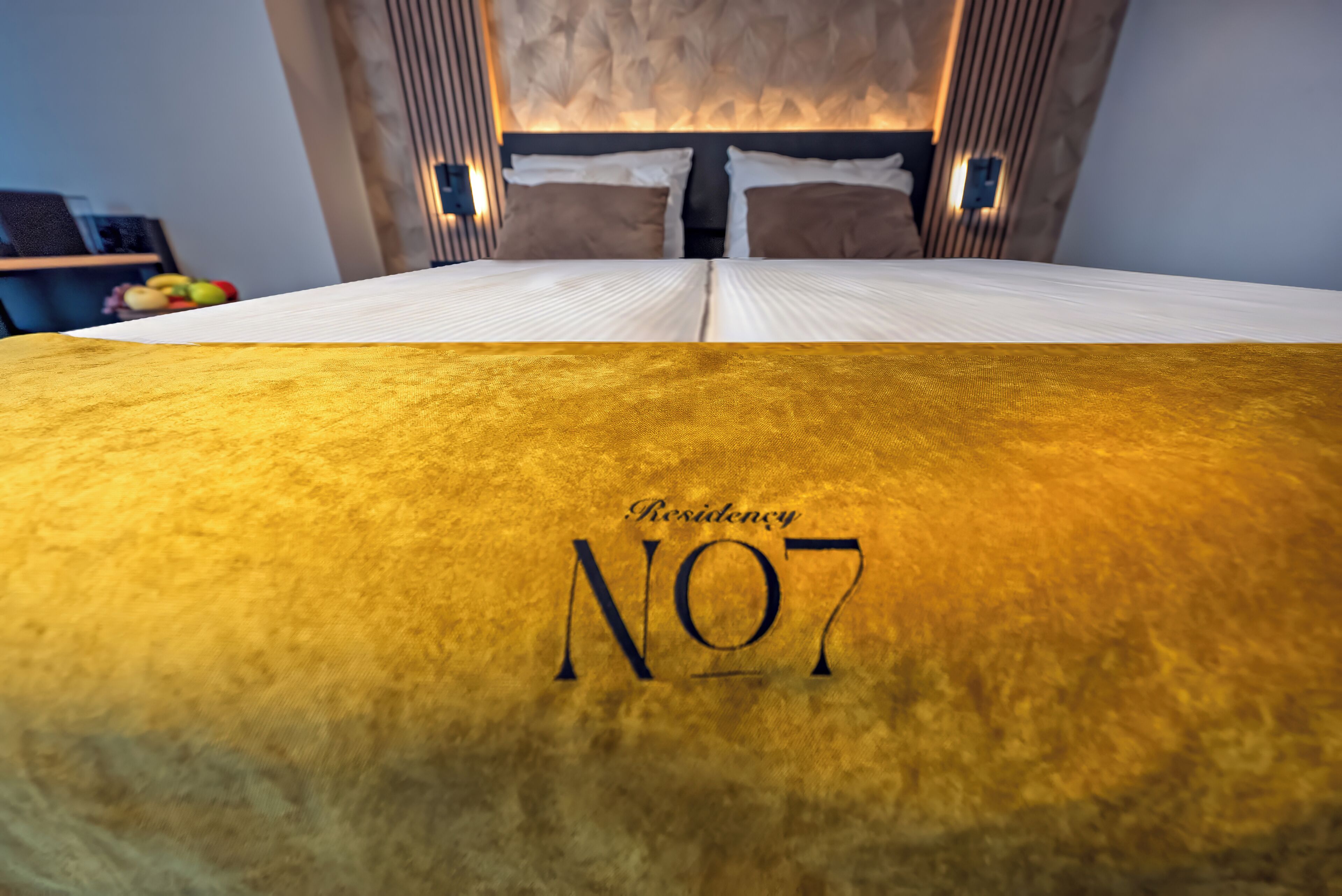 A hotel room with a bed covered in a golden blanket, a headboard with built-in lighting, and a visible wall-mounted shelf with fruits.