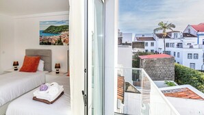 Hotelkamer met twee bedden, een schilderij van een kustlandschap en een balkon met uitzicht op de stad.