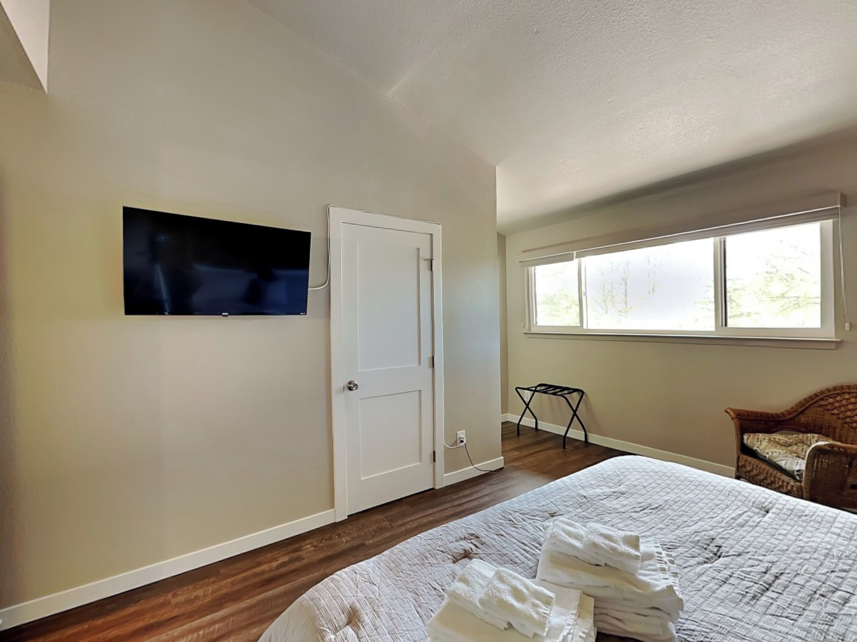 Un dormitorio con cama, televisión montada en la pared, puerta, ventana y una silla.