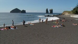 A beach with people sunbathing and swimming, rocky formations in the background, and a clear sky.