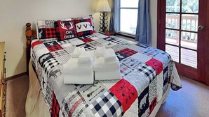 A bedroom with a large bed, a nightstand, a lamp, a window with curtains, and a door with glass panels. - Pinon Park A15 by Ruidoso Vacation Rentals (Ruidoso)