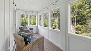 A covered porch with wicker furniture and large windows offering a view of a forest.