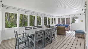 A covered outdoor dining area with a wooden table and chairs, large windows, and a view of the forest.