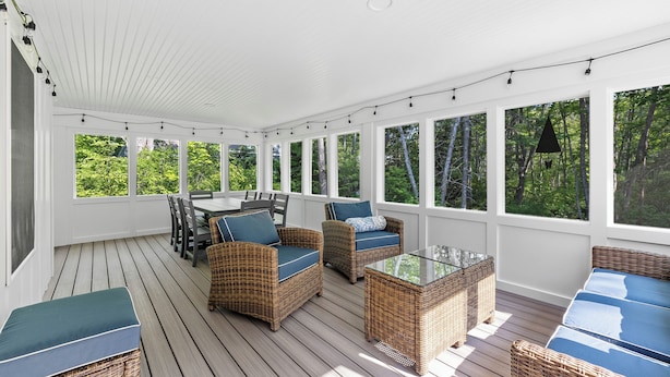 A covered outdoor seating area with wicker furniture and glass tables, surrounded by large windows offering a view of the greenery outside.