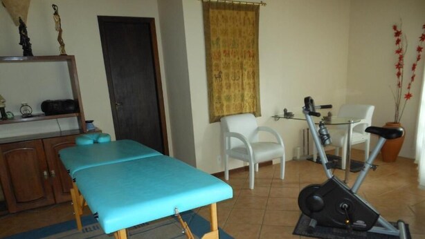 Een kamer met een massagetafel, een hometrainer en een kleine witte stoel.