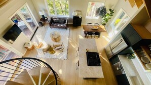 A spacious living area with a staircase, a dining table with chairs, a kitchen area, and large windows providing natural light.