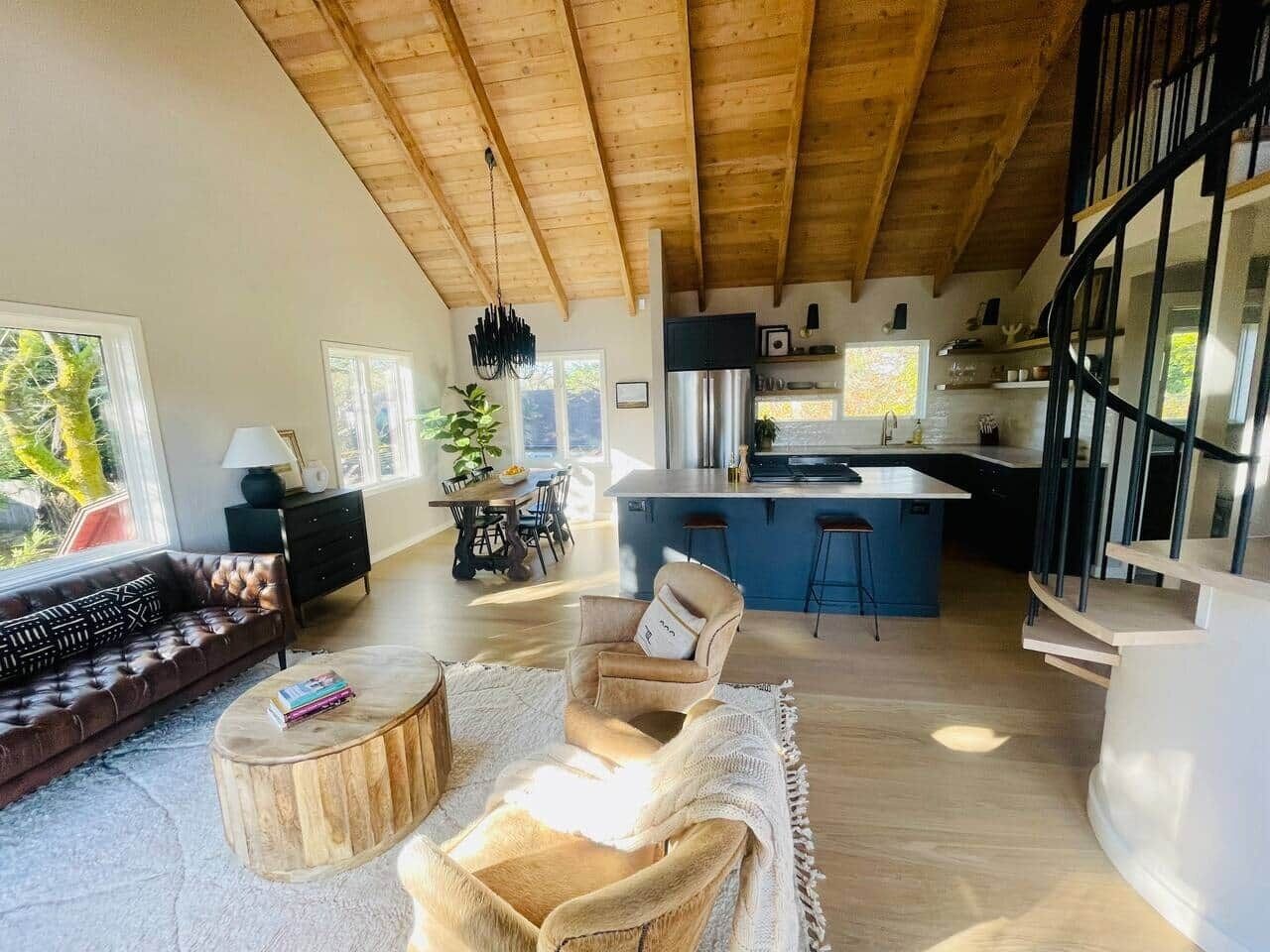 A spacious living area with a wooden ceiling, a sofa, a coffee table, and a chair. There is a kitchen area with a bar and a dining table in the background.