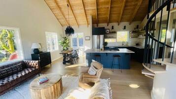 A spacious living area with a wooden ceiling, a sofa, a coffee table, and a chair. There is a kitchen area with a bar and a dining table in the background.