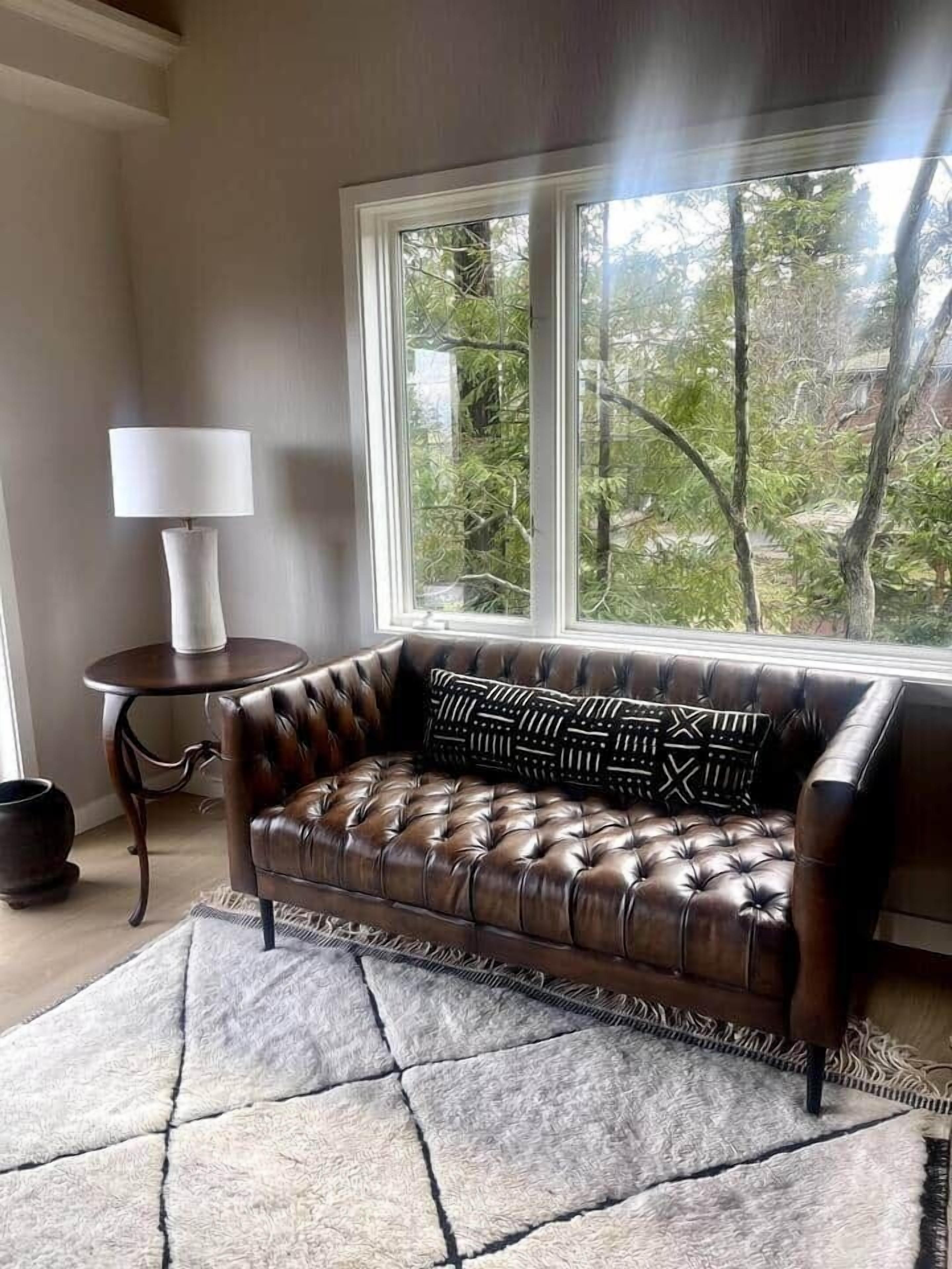 Un canapé en cuir agrémenté de coussins à motifs, une petite table ronde avec une lampe, et une vue sur les arbres par une grande fenêtre.