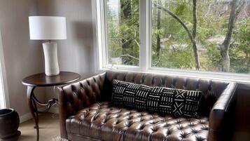 Un canapé en cuir agrémenté de coussins à motifs, une petite table ronde avec une lampe, et une vue sur les arbres par une grande fenêtre.
