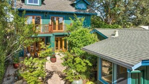 A two-story house with a balcony, surrounded by lush greenery and a stone patio.