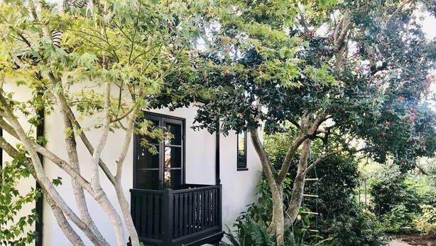 Un bâtiment blanc doté d’un balcon noir, entouré d’une végétation luxuriante et d’arbres.