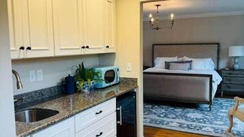 A compact hotel room with a kitchenette, a bed with a headboard, and a small seating area.