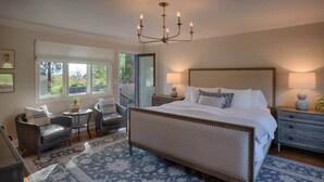 Une chambre à coucher avec un grand lit, deux tables de chevet avec des lampes, un lustre, une chaise, une petite table et une fenêtre donnant sur un paysage verdoyant.