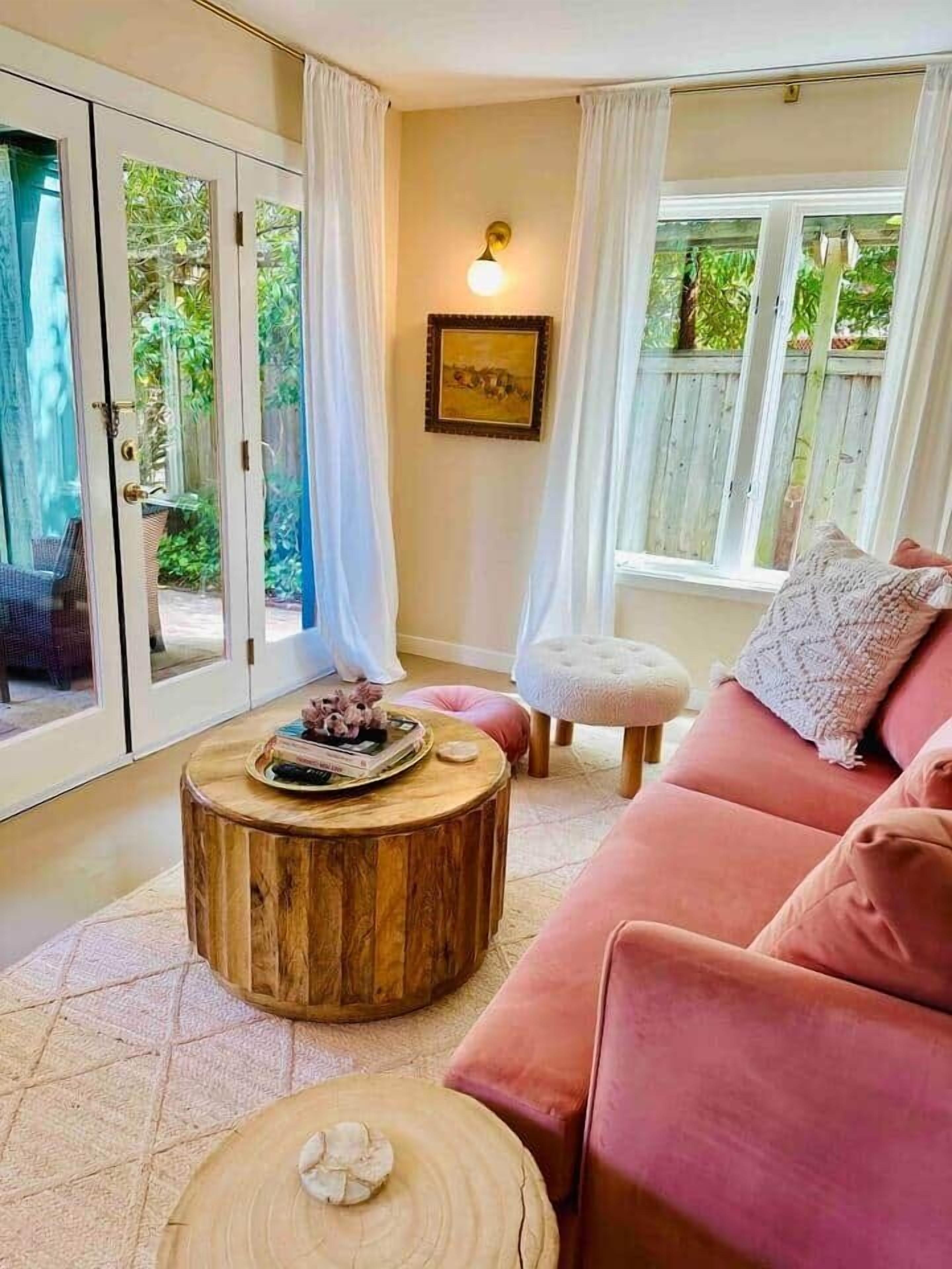 A cozy living room with a pink sofa, a wooden coffee table, and a view of a garden through glass doors.