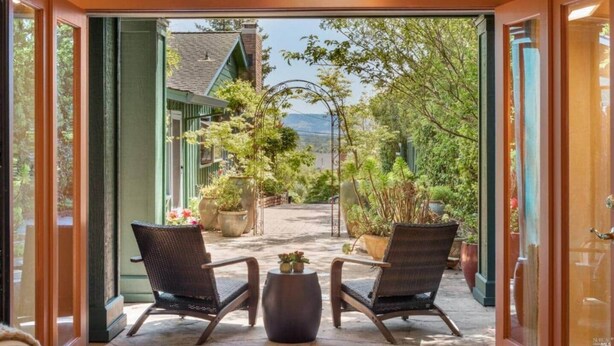 Une porte ouverte donne sur un patio où se trouvent deux fauteuils en osier et une petite table, le tout entouré de plantes en pot et offrant une vue sur un bâtiment verdoyant et des arbres.
