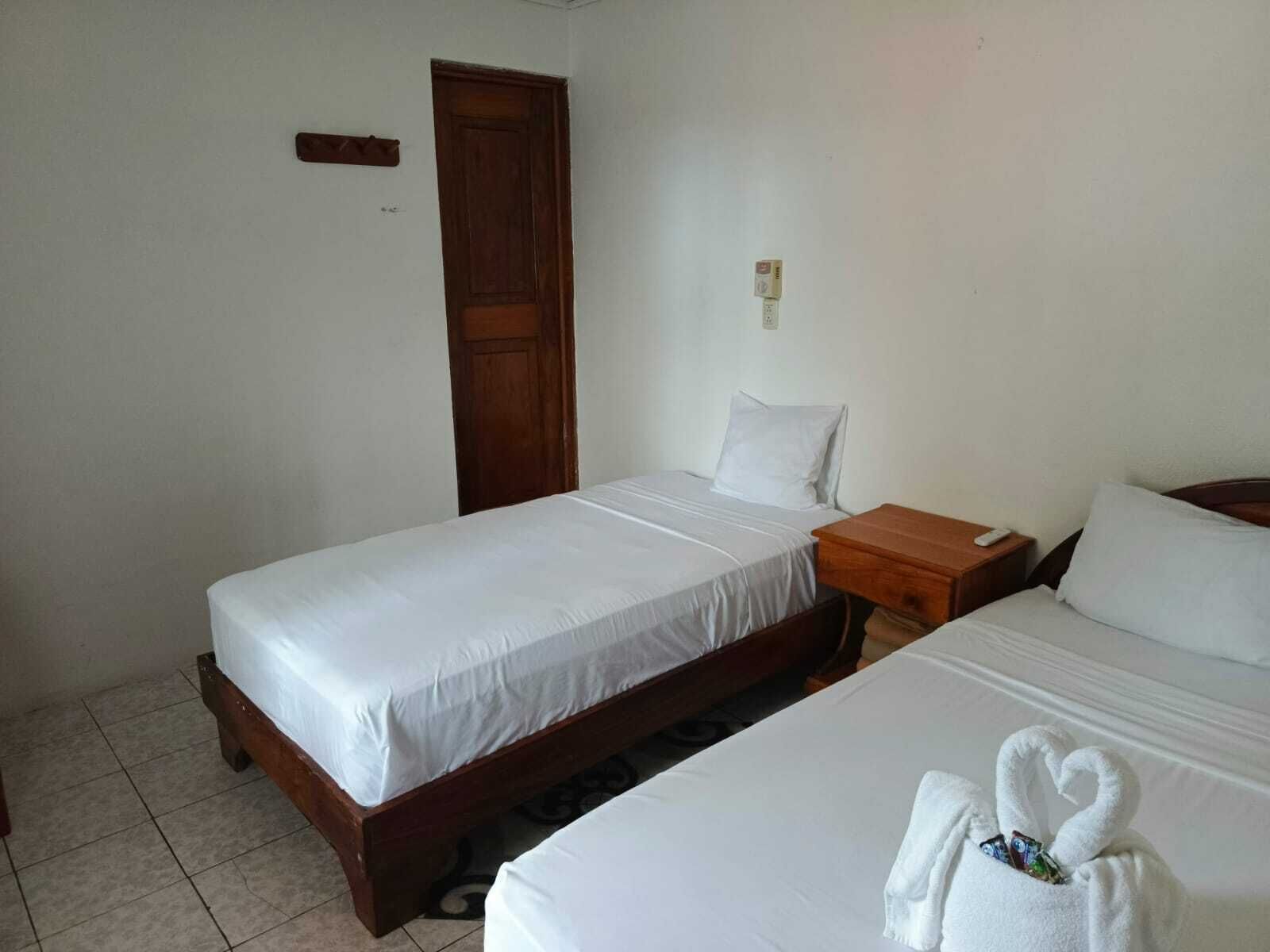 A simple, clean room with two beds, a wooden door, and a small bedside table with a remote control.