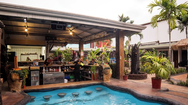 A pool area with a bar and seating, surrounded by potted plants and palm trees. - Arenal Hostel Resort (La Fortuna)