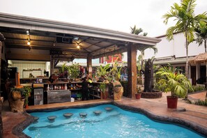 A pool area with a bar and seating, surrounded by potted plants and palm trees. - Arenal Hostel Resort (La Fortuna)