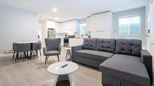 Een moderne woonkamer met een grijze hoekbank, een witte salontafel en twee grijze fauteuils. Achtergrond: een keuken met witte kasten en een raam met jaloezieën.