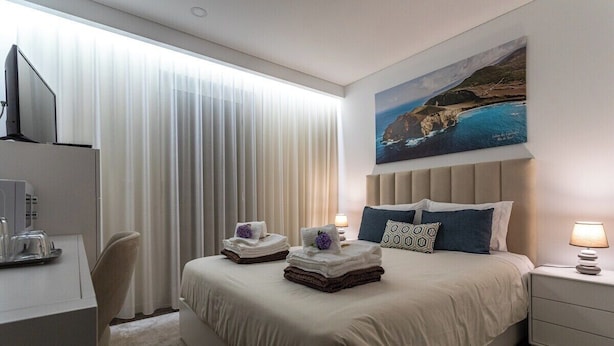 Ein Hotelzimmer mit einem Bett, einem Schreibtisch, einem Stuhl, einem Fernseher, einem Gemälde einer Küstenlandschaft und einem fenster mit Vorhängen.