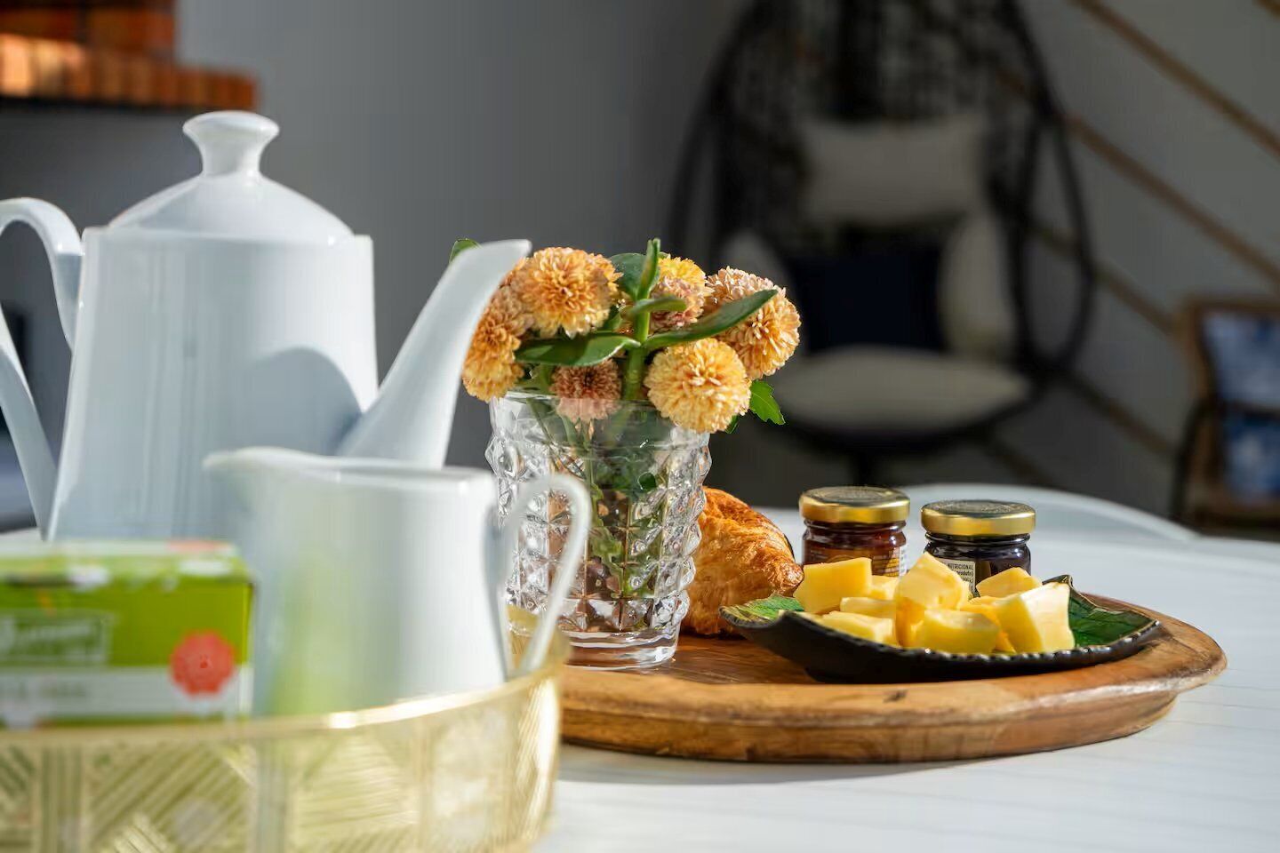 Une table dressée pour le petit-déjeuner avec une théière, un verre de jus de fruits, une assiette de fromage et un vase de fleurs.