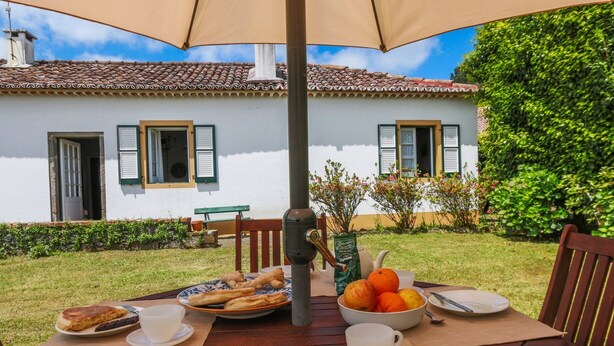 An outdoor dining table with food and drinks, a patio umbrella, and a white building with green shutters in the background.