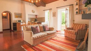 A spacious living area with a sofa, armchairs, and a wooden ceiling. A kitchen area is visible in the background.