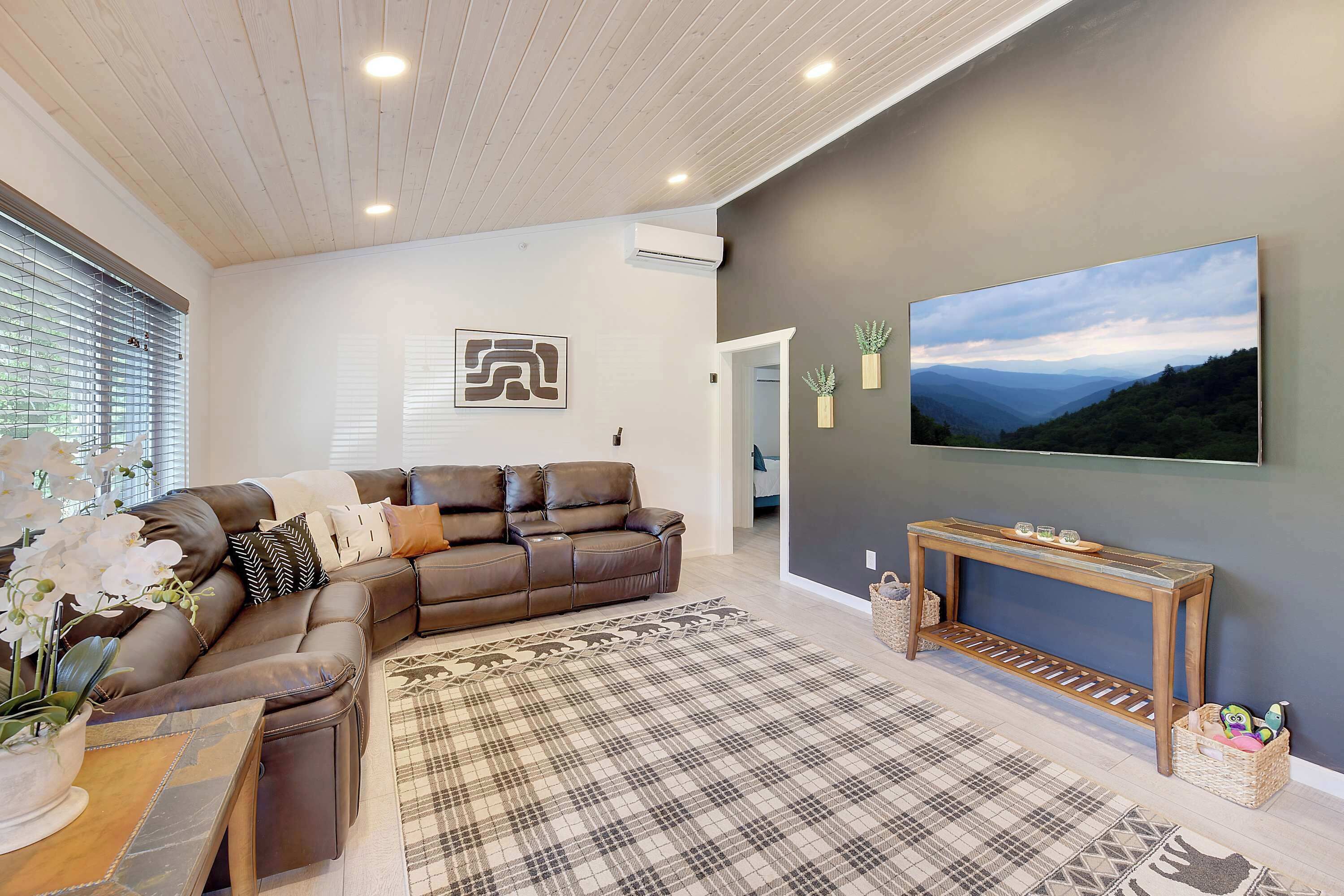 A modern living room with a brown leather sofa, a plaid rug, a wooden console table, and a large landscape TV on the wall.