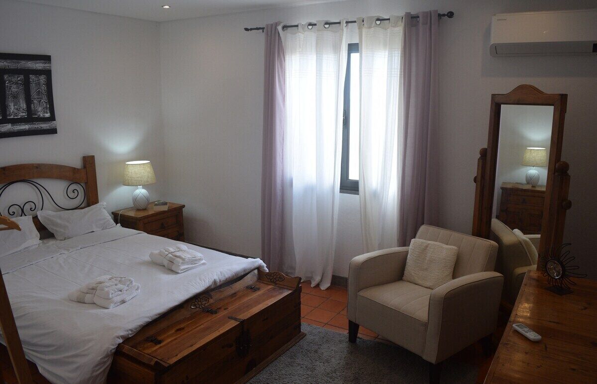 Ein Schlafzimmer mit Bett, Nachttischen, einem Sessel, einem Spiegel und einem Fenster mit Vorhängen.