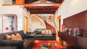 A spacious living room with a leather sofa, a glass coffee table, and a red rug. A staircase with a wooden railing leads to an upper level. - GuestReady - Casa da Boa Viagem (Ribeira Grande)
