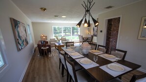 A dining area with a long wooden table set for six, surrounded by chairs. There is a chandelier above the table, and a large window provides natural light. - Luxurious, fully renovated 5 bedroom home great indoor & outdoor living spaces (Newton)