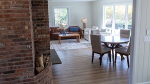 A spacious living area with a brick fireplace, a wooden coffee table, and a dining table with chairs. There are large windows providing natural light and a view of the outdoors. - Luxurious, fully renovated 5 bedroom home great indoor & outdoor living spaces (Newton)