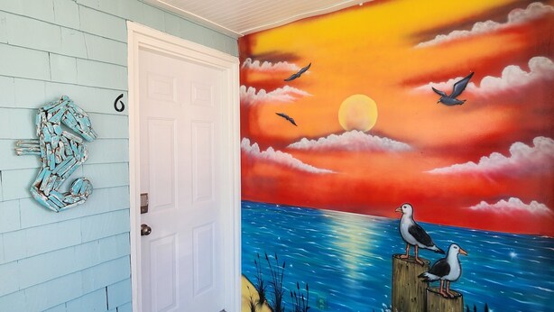 Une pièce ornée d’une fresque représentant un coucher de soleil sur l’océan, dotée d’un plancher en bois et d’une porte sur laquelle est affiché le numéro « 6 ».