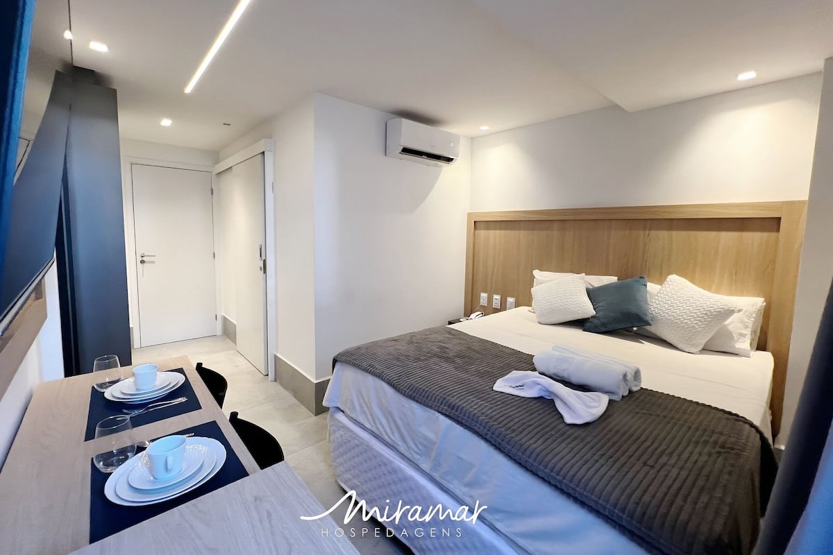 A hotel room with a neatly made bed, a dining table set with plates and glasses, and an air conditioning unit on the wall.