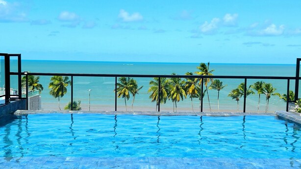 A pool with clear blue water, a metal railing, and a view of the ocean and palm trees. - Legacy Suites (Joao Pessoa)