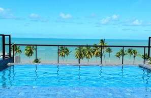 A pool with clear blue water, a metal railing, and a view of the ocean and palm trees. - Legacy Suites (Joao Pessoa)