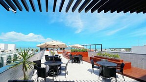A rooftop terrace with outdoor seating, umbrellas, and a view of the sea. - Legacy Suites (Joao Pessoa)