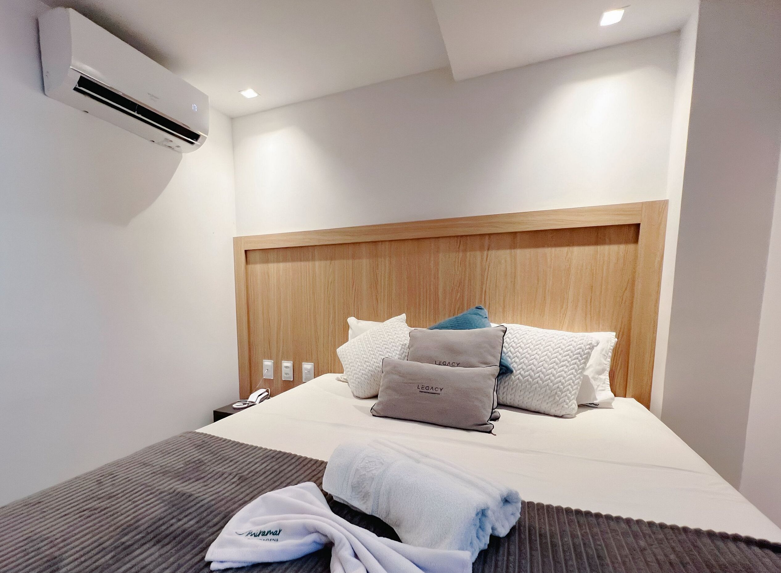 Une chambre d’hôtel avec une tête de lit en bois, un lit avec une literie blanche et plusieurs oreillers, une table de chevet avec un téléphone et une lampe, et un appareil de climatisation fixé au mur.
