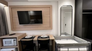 A hotel room with a flat-screen TV mounted on a wooden panel, a crib with a brand name, a microwave, and a dining area with chairs and a table set for two. - Legacy Suites (Joao Pessoa)