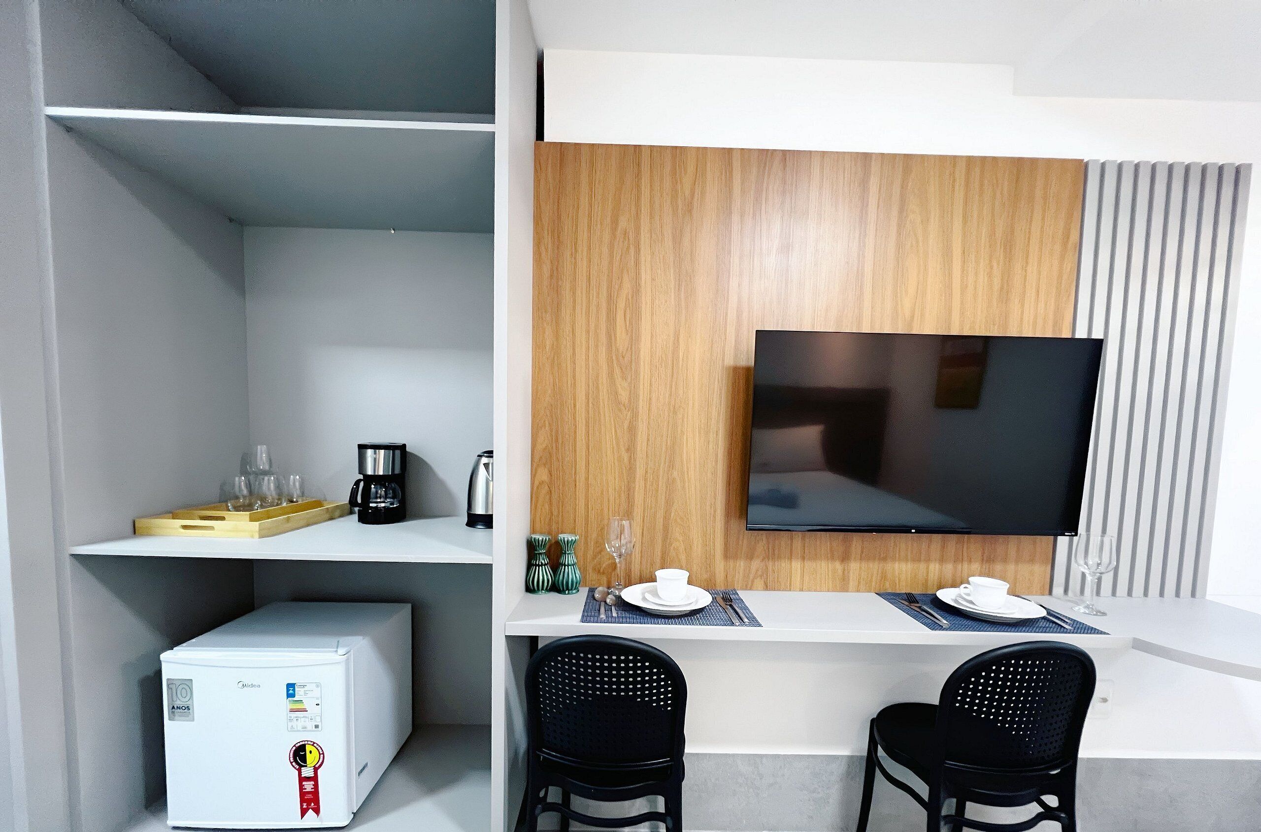 Une chambre d’hôtel moderne avec un bureau blanc, deux chaises noires, un téléviseur à écran plat fixé à un mur en bois, un mini-réfrigérateur et une étagère avec des verres et une machine à café.