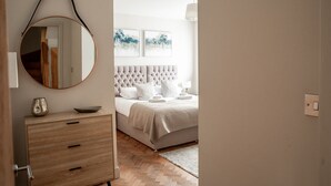A bedroom with a tufted headboard, a round mirror, a wooden dresser, and a bed with white bedding.
