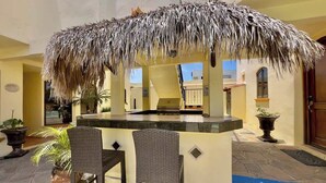 An outdoor bar area with a thatched roof, two wicker chairs, and potted plants.