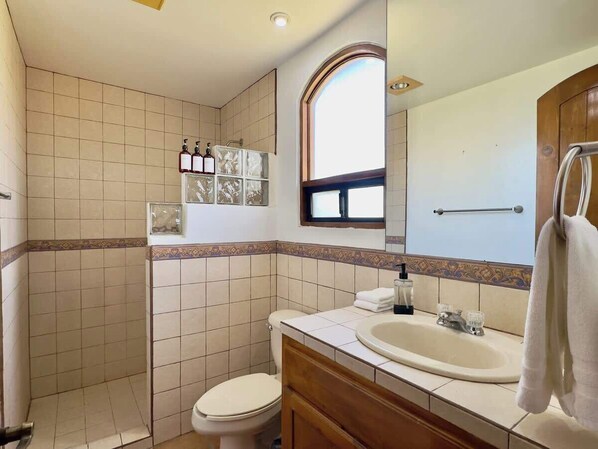 Baño con inodoro, lavamanos y ducha. Hay una ventana sobre el lavamanos y un espejo en la pared.