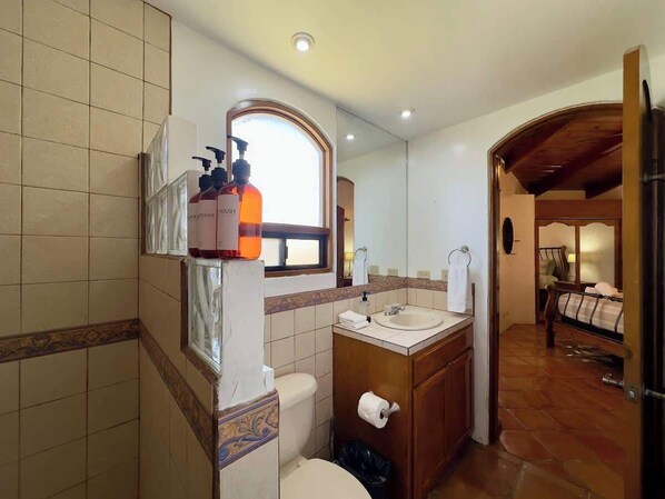 Un baño con inodoro, lavamanos y zona de ducha. Se ve una ventana con vistas al exterior.